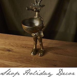 May include: Gold metal deer figurine holding a bowl. The deer is wearing a jacket and has a pipe in its mouth. The figurine is a holiday decor item.