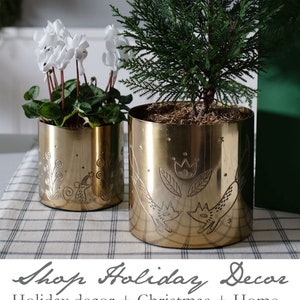 May include: Two gold metal planters with a decorative design. The larger planter has a design of two foxes and a flower. The smaller planter has a design of a mouse and a flower. Both planters are on a checkered tablecloth.