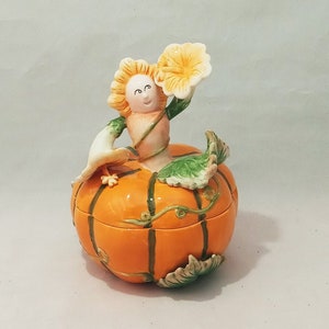 Puede incluir: Una caja de cerámica en forma de calabaza con una figurita caprichosa, sonriente, de pelo verde en la parte superior. La figurita sostiene una flor amarilla y tiene un pájaro blanco posado en su brazo.