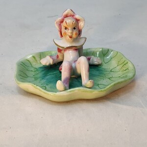 Puede incluir: Una figurita de cerámica de un elfo fantasioso sentado en un plato verde en forma de hoja. El elfo lleva un atuendo a rayas rosa y blanco con un collar dorado y un sombrero de flor rojo y blanco.