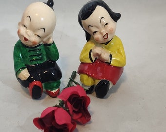 Könnte beinhalten: Zwei kleine Keramikfiguren von Kindern in traditioneller chinesischer Kleidung. Der Junge trägt ein grünes und schwarzes Outfit und das Mädchen trägt ein gelbes und rotes Outfit. Beide lächeln und halten sich an den Händen. Vor ihnen liegen zwei rote Rosen.