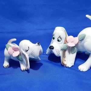 May include: Two white porcelain figurines of dogs with pink flowers and green leaves. The dogs are in a playful pose, with one dog looking at the other.