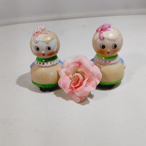 May include: Two ceramic dolls with painted faces, wearing blue and green dresses, with pink bows in their hair. The dolls are standing on a white surface with a pink rose in front of them.