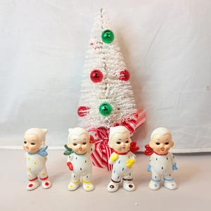 Può includere: Quattro statuine natalizie vintage in ceramica di neonati in tutine bianche con pois e accenti colorati. Un piccolo albero di Natale bianco con palline rosse e verdi e un fiocco a righe rosse e bianche è dietro le statuine.