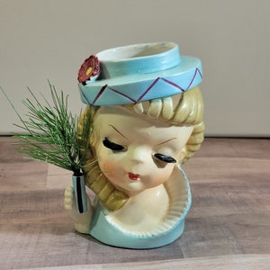 Może przedstawiać: Wazon w kształcie głowy z ceramiki w stylu vintage przedstawiający kobietę z blond włosami, w jasnoniebieskim kapeluszu z bordowym kwiatem i pasującą sukienką. Wazon zawiera gałązkę zielonych liści. Twarz ma malowane cechy.
