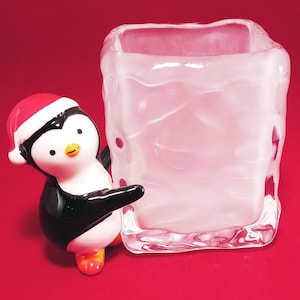 Puede incluir: Una figurita de pingüino de cerámica con un gorro de Papá Noel rojo, sosteniendo un jarrón cuadrado de vidrio esmerilado. El pingüino es negro y blanco con pies naranjas.