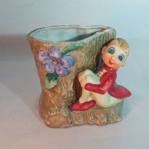 Könnte beinhalten: Ein Keramik-Pflanztopf in Form eines Baumstamms mit einer kleinen Elfenfigur, die darauf sitzt. Der Elf trägt eine rote Jacke und eine weiße Hose. Der Topf ist mit einer lila Blume bemalt.