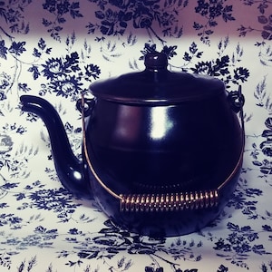 Puede incluir: Una tetera negra con asa dorada y tapa de resorte. La tetera está sobre un fondo blanco con un patrón floral.