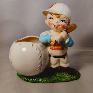 May include: Ceramic figurine of a baseball player holding a bat. The player is wearing a blue and white uniform with the words "The Slugger" on the front. The figurine is holding a white baseball with a red stitching design.