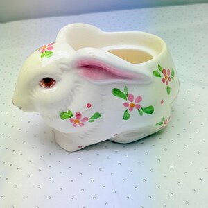 May include: White ceramic bunny-shaped planter with pink-lined ears and brown eyes. The planter is decorated with pink and green floral accents. A charming Easter or spring-themed decorative item.
