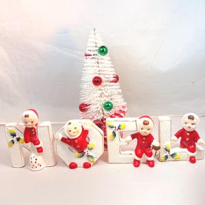 May include: A festive Christmas display featuring the word "NOEL" spelled out with white ceramic letters. Each letter has a small figurine of a child in a red suit and hat. A small white Christmas tree with red and green ornaments is in the background.