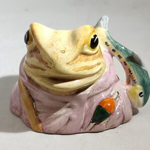 May include: A ceramic frog-shaped mug with a fish handle. The frog is light yellow with brown stripes and black eyes. It wears a pink shirt with a fishing float. The fish handle is green, blue, and yellow.