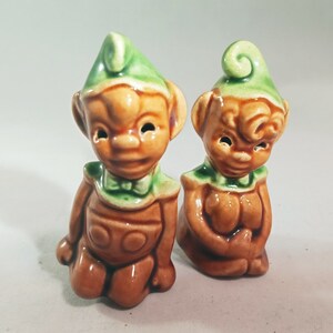 May include: A pair of ceramic elf salt and pepper shakers. The shakers are brown with green hats and are sitting on a white surface.