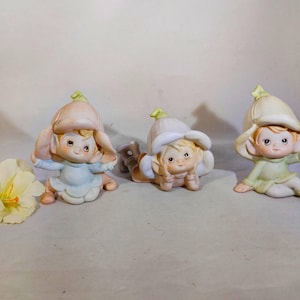 Puede incluir: Tres figuritas de cerámica de elfos con sombreros con forma de flor. Los elfos llevan atuendos de diferentes colores y tienen rasgos faciales pintados.