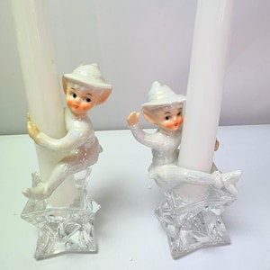 May include: Two ceramic elf candle holders with an iridescent finish, each featuring a small elf figure climbing a tall white taper candle. The figures are seated on clear, star-shaped glass bases with faceted edges and a transparent, crystalline appearance.