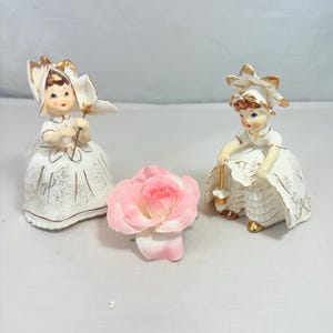 Könnte beinhalten: Zwei Vintage-Porzellanfiguren von Mädchen in weißen Kleidern mit goldenen Akzenten. Eine hält eine Blume, die andere eine Laterne. Zwischen ihnen liegt eine rosa Rose. Die Figuren haben detaillierte Gesichter und stehen auf einer weißen Oberfläche.