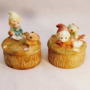 Puede incluir: Dos cajas de cerámica para guardar pequeños objetos con figuras de hadas o elfos de fantasía. Una caja presenta un hada sentada con un hongo, mientras que la otra presenta un hada tumbada con un hongo. Ambas cajas están decoradas con un patrón de veta de madera.