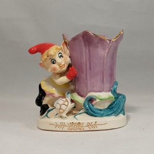 Könnte beinhalten: Eine Keramikfigur eines Zwergs, der einen violetten Blumentopf hält. Der Zwerg trägt einen roten Hut und ein weiß-gelbes Kleid. Eine kleine Schildkröte sitzt auf dem Boden neben dem Zwerg.