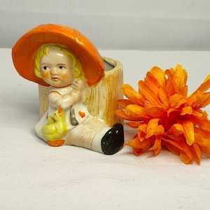 May include: A vintage ceramic planter featuring a young girl with blonde hair wearing an orange hat, holding a yellow duckling. The planter is cream-colored with a tree trunk design. An orange flower is next to the planter.