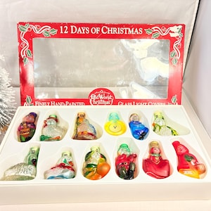 May include: A set of twelve hand-painted glass Christmas ornaments, each depicting a different character from the "12 Days of Christmas" carol. The ornaments are displayed in a white box with a red lid that reads "12 Days of Christmas".