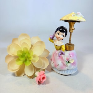 May include: A vintage porcelain figurine of a woman in a yellow and pink dress, holding her hair, with a yellow parasol. A faux succulent and a pink rose are next to the figurine.