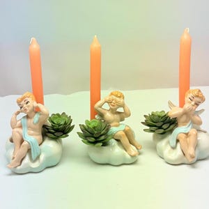 Puede incluir: Tres candelabros de cerámica con figuras de querubines sentados en nubes, cada uno sosteniendo una planta suculenta. Altas velas naranjas se insertan en la espalda de los querubines. Los querubines están en varias poses, uno cubriéndose los ojos.