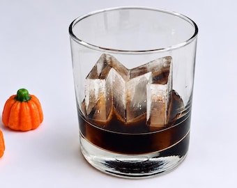 Könnte beinhalten: Ein klares Glas mit dunkler Flüssigkeit und einem großen, kantigen Eiswürfel, begleitet von drei kleinen orangefarbenen kürbisförmigen Objekten mit grünen Stielen. Der Text SHACKLETON ICE und *Garnish Optional sind im Bild zu sehen.