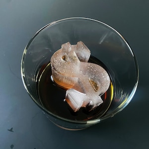 Puede incluir: Un vaso con un líquido oscuro y un cubito de hielo con forma de signo de dólar.