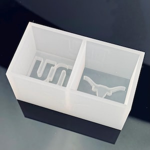 Op de afbeelding: Een heldere, rechthoekige harsvorm met twee compartimenten. Eén compartiment heeft de letters "UN" en de andere een longhorn-silhouet. De basis van de vorm is zwart.