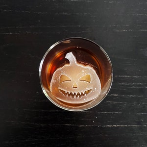 Op de afbeelding: Een glas met een donkere vloeistof en een heldere ijsblokje in de vorm van een Halloween-pompoen. Het ijsblokje heeft een gesneden gezicht met een brede glimlach en twee ogen.