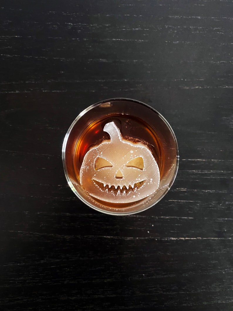 Op de afbeelding: Een glas met een donkere vloeistof en een heldere ijsblokje in de vorm van een Halloween-pompoen. Het ijsblokje heeft een gesneden gezicht met een brede glimlach en twee ogen.