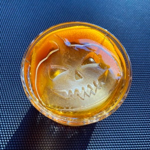 Op de afbeelding: Een glas gevuld met een gele vloeistof en een grote ijsblokje in de vorm van een jack-o'-lantern.
