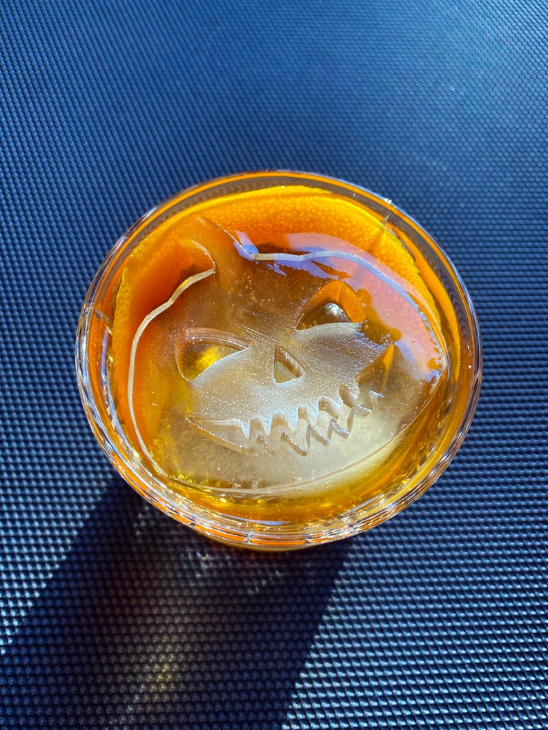 Op de afbeelding: Een glas gevuld met een gele vloeistof en een grote ijsblokje in de vorm van een jack-o'-lantern.