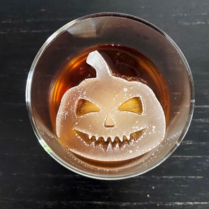 Op de afbeelding: Een helder glas met een bruine vloeistof en een witte ijsblokje in de vorm van een jack-o'-lantern.