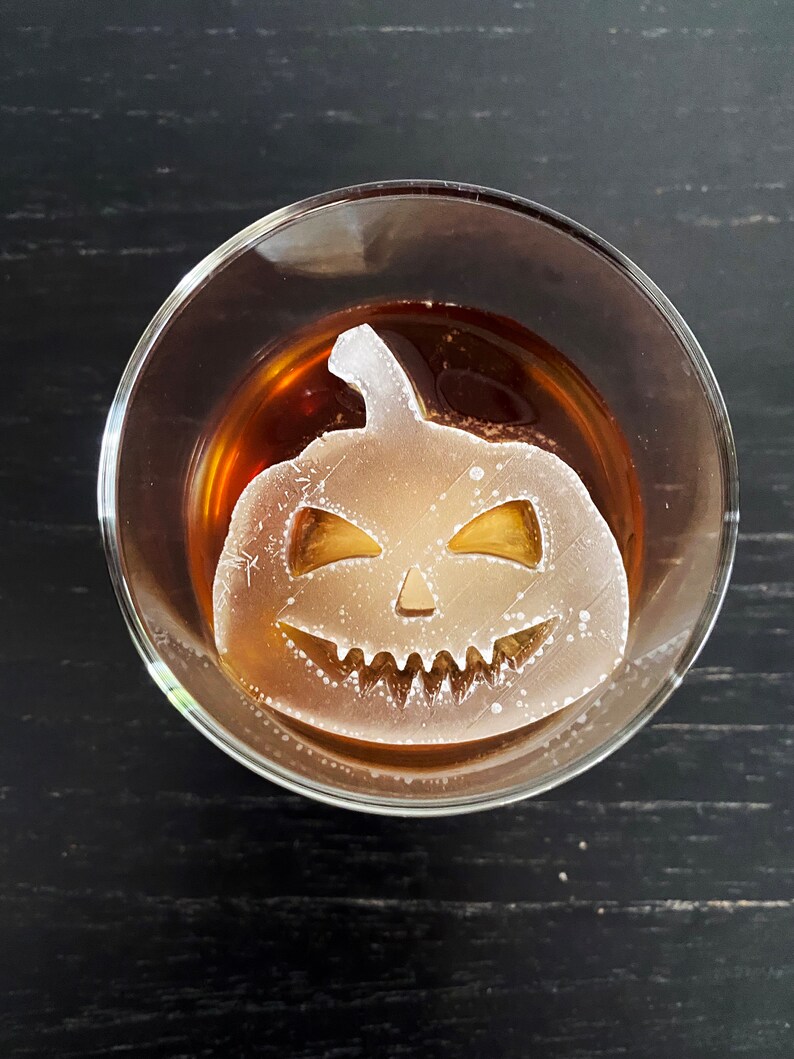 Op de afbeelding: Een helder glas met een bruine vloeistof en een witte ijsblokje in de vorm van een jack-o'-lantern.