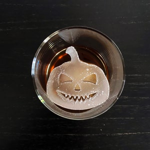Op de afbeelding: Een helder glas met een donkerbruine vloeistof en een witte ijsblokje in de vorm van een jack-o'-lantern.