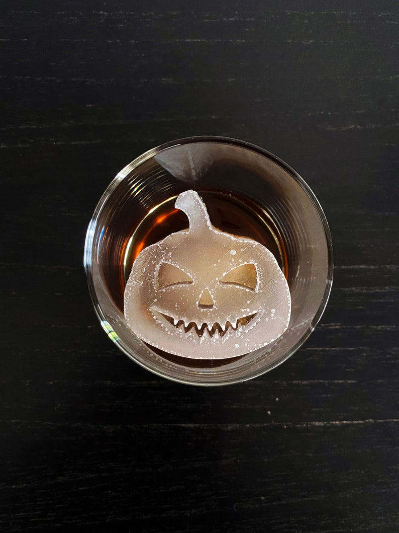 Op de afbeelding: Een helder glas met een donkerbruine vloeistof en een witte ijsblokje in de vorm van een jack-o'-lantern.