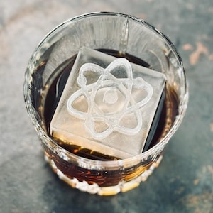 May include: A clear, etched ice cube in a rocks glass filled with a dark amber liquid. The ice cube features a detailed atom symbol design. The glass has a textured base and is set against a blurred background.