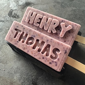 Może przedstawiać: Dwa prostokątne, jasnofioletowe lody na patyku z wytłoczonymi imionami "HENRY" i "THOMAS". Lody mają widoczne plamki i są na drewnianych patyczkach. Lody spoczywają na czarnej powierzchni.