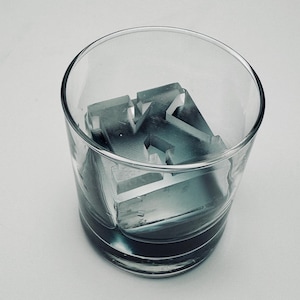 Puede incluir: Un vaso transparente lleno de un objeto gris parecido a hielo con forma de la letra "H". El hielo está parcialmente sumergido en un líquido oscuro, creando una estética fresca. El vaso está sobre un fondo blanco.