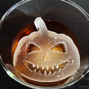 Op de afbeelding: Een glazen glas met een bruine vloeistof en een witte plastic pompoen-ijsblokje. Het ijsblokje heeft een gesneden jack-o'-lantern gezicht.