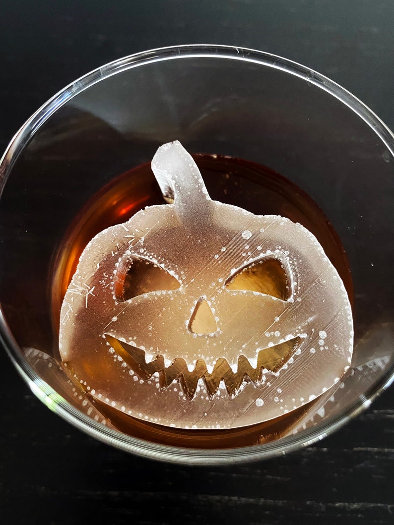 Op de afbeelding: Een glazen glas met een bruine vloeistof en een witte plastic pompoen-ijsblokje. Het ijsblokje heeft een gesneden jack-o'-lantern gezicht.