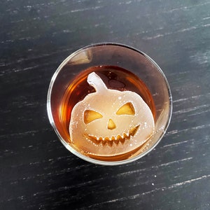 Op de afbeelding: Een helder glas met een bruine vloeistof en een witte ijsblokje in de vorm van een jack-o'-lantern.