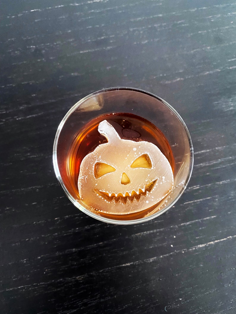 Op de afbeelding: Een helder glas met een bruine vloeistof en een witte ijsblokje in de vorm van een jack-o'-lantern.