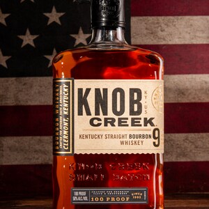 May include: A clear glass bottle of Knob Creek 9-year Kentucky Straight Bourbon Whiskey. The bottle has a brown label with gold lettering and a black cap. The bottle is sitting on a wooden surface in front of an American flag.