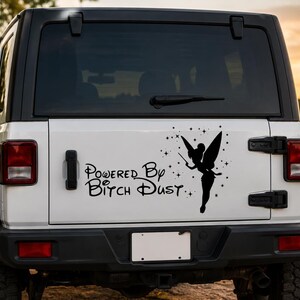 May include: A black vinyl decal applied to the rear window of a white vehicle. The decal features a silhouette of a fairy with wings and a wand, surrounded by small stars, and the text "Powered By Bitch Dust" in a stylized cursive font.