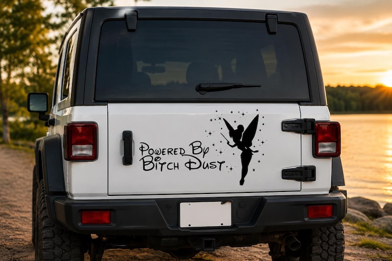 May include: A black vinyl decal applied to the rear window of a white vehicle. The decal features a silhouette of a fairy with wings and a wand, surrounded by small stars, and the text "Powered By Bitch Dust" in a stylized cursive font.