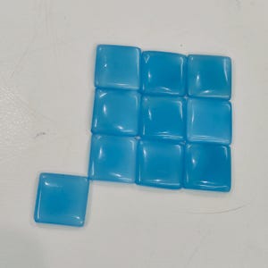 May include: A collection of twelve square, light blue gemstones. Eleven are arranged in a grid, with one gemstone slightly offset. The stones have a smooth, polished surface and a translucent quality, reflecting light.