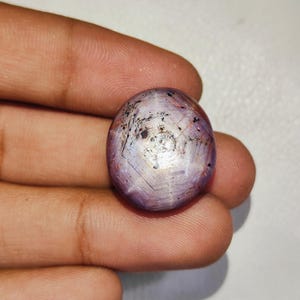 May include: A round, cabochon-cut star sapphire gemstone featuring a translucent, light purple and pink hue. The stone displays a distinct six-rayed star effect across its surface, with visible dark inclusions and natural internal striations.