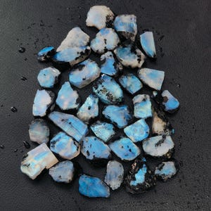 May include: A collection of raw, uncut moonstone gemstones. The stones display a mix of colors, including white, blue, and black. The gemstones are irregularly shaped and vary in size, with a polished, reflective surface. The background is a dark, textured surface.